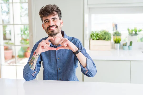 Young man wearing casual shirt sitting on white table smiling in love showing heart symbol and shape with hands. Romantic concept.