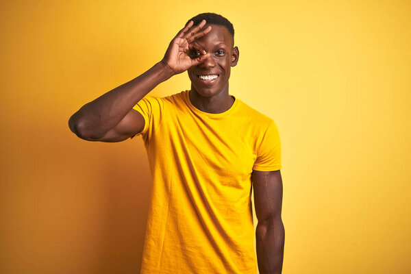 Young african american man wearing casual t-shirt standing over isolated yellow background doing ok gesture with hand smiling, eye looking through fingers with happy face.
