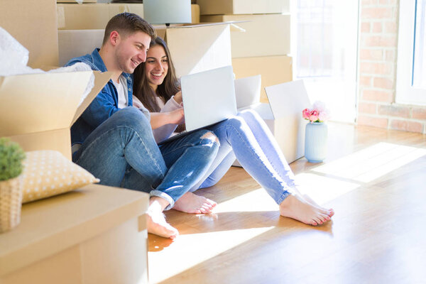 Young couple sitting on the floor of new house around cardboard boxes using laptop, smiling happy for new apartment