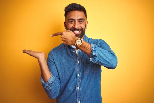 独立した黄色の背景に立ってデニムシャツを着ている若いインド人男性は 手で提示しながら カメラに驚いて笑顔と指で指して — ストック写真