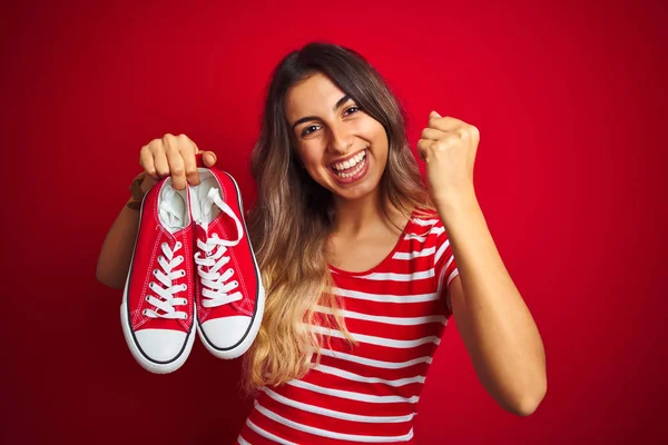 Joven Hermosa Mujer Sosteniendo Zapatillas Sobre Fondo Rojo Aislado Gritando —  Fotos de Stock