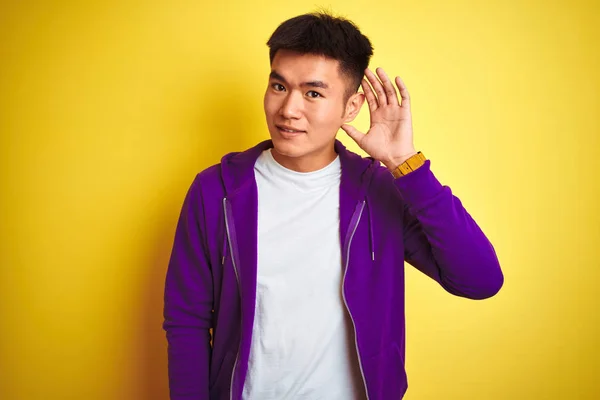 噂やゴシップに耳を傾ける耳の上に手で笑みを浮かべて孤立した黄色の背景の上に立って紫色のスウェットシャツを着て若いアジアの中国人男性 病気の概念 — ストック写真