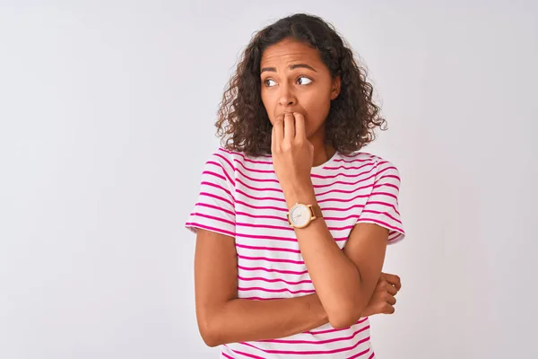 Young brazilian woman wearing pink striped t-shirt standing over isolated white background looking stressed and nervous with hands on mouth biting nails. Anxiety problem.