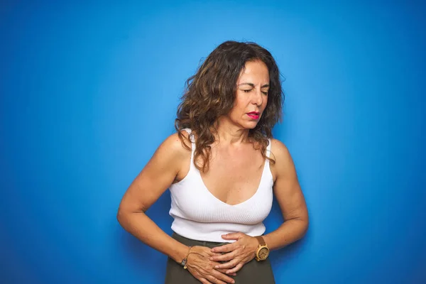 Middle age senior woman with curly hair standing over blue isolated background with hand on stomach because indigestion, painful illness feeling unwell. Ache concept.
