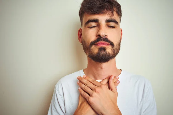 Young man with tattoo wearing t-shirt standing over isolated white background smiling with hands on chest with closed eyes and grateful gesture on face. Health concept.