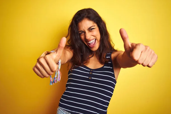 年轻的漂亮女人穿着条纹T恤站在孤立的黄色背景批准做积极的姿态与手 竖起大拇指微笑和高兴的成功 获胜者手势 — 图库照片
