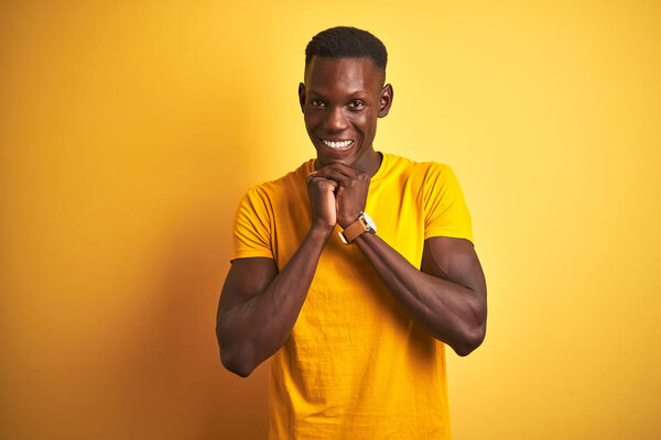 Young african american man wearing casual t-shirt standing over isolated yellow background laughing nervous and excited with hands on chin looking to the side