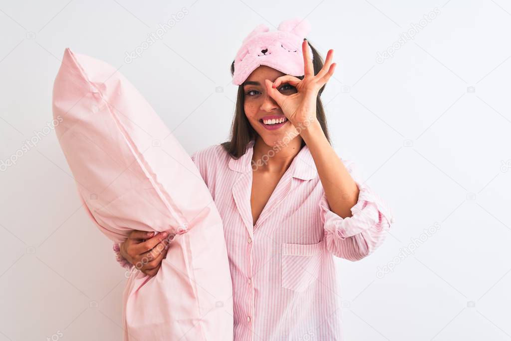 Beautiful woman wearing sleep mask pajama holding pillow over isolated white background with happy face smiling doing ok sign with hand on eye looking through fingers