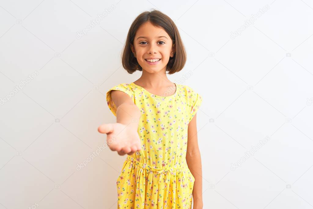 Young beautiful child girl wearing yellow floral dress standing over isolated white background smiling cheerful offering palm hand giving assistance and acceptance.
