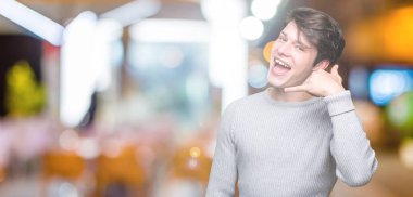 Genç yakışıklı adam giyen kış kazak yapıyorsun gülümseyen izole arka plan üzerinde telefon hareketi ile el ve parmak telefonda konuşuyor gibi. Kavramları iletişim.