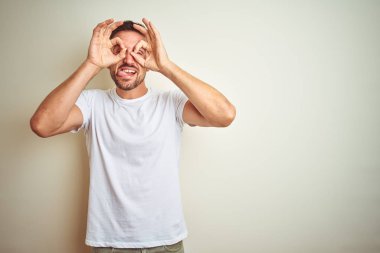 Dilini, parmaklarının arasından baktığını yapışmasını dürbün gibi ok jest yapıyor izole arka plan üzerinde rahat beyaz t-shirt giyen genç yakışıklı adam. Deli ifade.