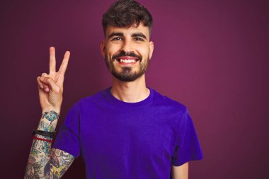 İzole edilmiş mor arka plan üzerinde duran dövme tişörtlü genç adam iki numaralı parmaklarıyla işaret ederken kendinden emin ve mutlu gülümsüyor..
