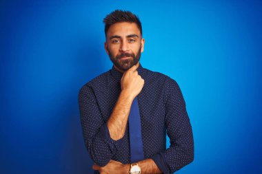 Zarif gömlek ve kravat giyen genç Hintli işadamı izole edilmiş mavi arka plan üzerinde duruyor kameraya kollarını kavuşturup gülümseyerek bakıyor ve çenesini kaldırıyor. Olumlu düşünüyorum..