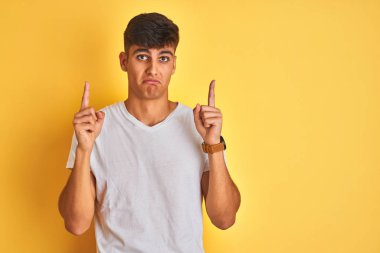 İzole edilmiş sarı arka plan üzerinde duran beyaz tişörtlü genç Hintli adam üzgün ve üzgün görünüyor, parmaklarıyla yönü gösteriyor, mutsuz ve depresif..