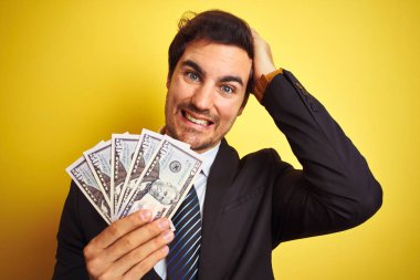 Takım elbiseli genç yakışıklı işadamı, dolar tutarak izole edilmiş sarı arka planda, elleri başının üstünde, utanç ve şaşkınlık dolu, sinirli ve hayal kırıklığına uğramış bir halde. Hata için korku ve üzüntü.