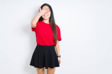 Güzel esmer kadın, kırmızı tişört ve etek giyerek izole arka planda gülümsüyor ve elleri yüzünde şaşkınlıktan gözleri kapayarak gülüyor. Kör kavram.