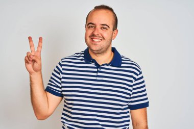 Sıradan çizgili polo giyen genç adam izole edilmiş beyaz arka planda durup kameraya bakıp zafer işareti yaparken gülümsüyor. İki numara..