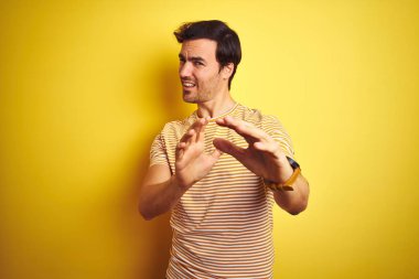 İzole edilmiş sarı arka planda, çizgili tişört giyen genç ve yakışıklı bir adam tiksinti reaksiyonu yüzünden tiksinti ve tiksinti ifadesi takınıyor. Eller havada. Can sıkıcı bir kavram..