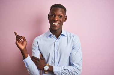 Mavi zarif gömlekli Afro-Amerikan bir adam pembe arka planda duruyor yüzünde kocaman bir gülümseme, eli ve parmağı kameraya bakıyor..