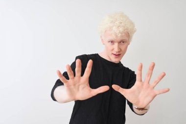 Siyah tişörtlü genç albino bir adam beyaz arka plan üzerinde duruyor korku ifadesinden korkuyor ve dehşete düşüyor. El hareketlerini durdur, şok içinde bağır. Panik konsepti.