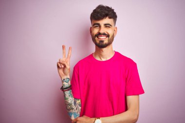İzole edilmiş pembe arka plan üzerinde duran tişörtlü genç adam kameraya bakıp gülümsüyor ve zafer işareti yapıyor. İki numara..