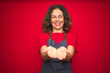 Kırmızı izole edilmiş arka planda önlük giyen orta yaşlı bir kadın avuçlarını birbirine değdirirken ya da el hareketi yaparken gülümsüyor. Tutun ve koruyun