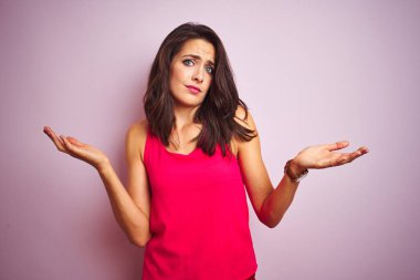 Genç güzel kadın giyen t-shirt üzerinde ayakta pembe izole arka plan clueless ve karıştı ifade ile silah ve eller kaldırdı. Şüphe kavramı.