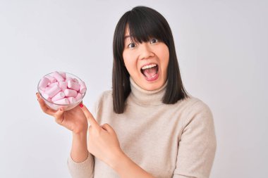 Güzel bir Çinli kadın, izole edilmiş beyaz arka planda şekerleme kasesiyle el ve parmağıyla işaret ederek çok mutlu olur.