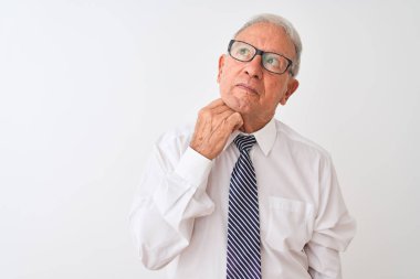 Kravat takan ve gözlük takan kıdemli gri saçlı işadamı tenha beyaz arka planda, elleri çenesinde soru sorarken, düşünceli bir ifade takınıyor. Düşünceli bir yüzle gülümsüyor. Şüphe kavramı.