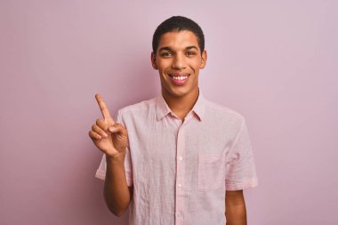 İzole edilmiş pembe arka planda dikilen rahat gömlekli genç, yakışıklı Arap adam bir numaralı parmağını gösterirken kendinden emin ve mutlu bir şekilde gülümsüyor..