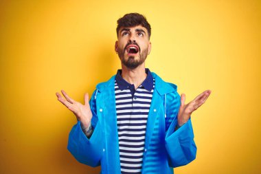 Yağmurluk giyen genç yakışıklı adam izole edilmiş sarı arka planda duruyor çılgın ve kızgın bir şekilde bağırıyor ve agresif bir ifade ve kolla bağırıyor. Hayal kırıklığı konsepti.