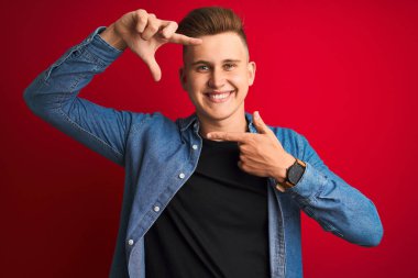 İzole edilmiş kırmızı arka plan üzerinde duran kot gömlekli yakışıklı bir adam gülümsüyor ve mutlu yüzlü elleri ve parmakları olan bir çerçeve yapıyor. Yaratıcılık ve fotoğrafçılık kavramı.