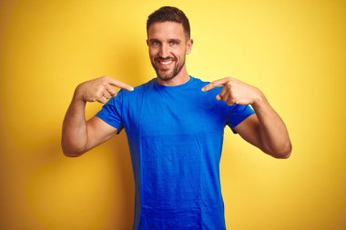Young handsome man wearing casual blue t-shirt over yellow isolated background looking confident with smile on face, pointing oneself with fingers proud and happy. clipart