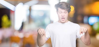 Casual beyaz t-shirt üzerinde izole giyen genç yakışıklı adam arka plan işaretleme aşağı reklam, sürpriz yüz ve açık ağız gösterilen parmakları ile