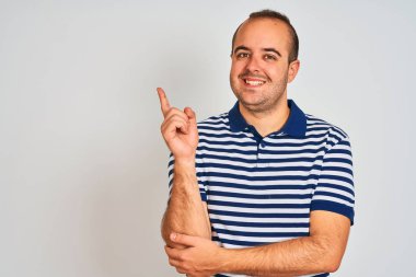 Sıradan çizgili polo giyen genç adam izole edilmiş beyaz arka planda duruyor. Yüzünde kocaman bir gülümseme var. Elinle kameraya bakıyor..