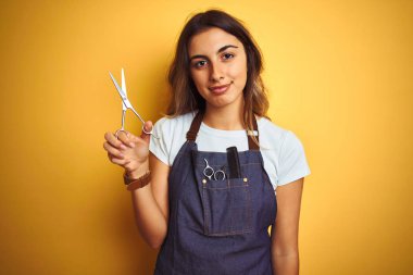 Young beautiful hairdresser woman holding scissors over yellow isolated background with a confident expression on smart face thinking serious