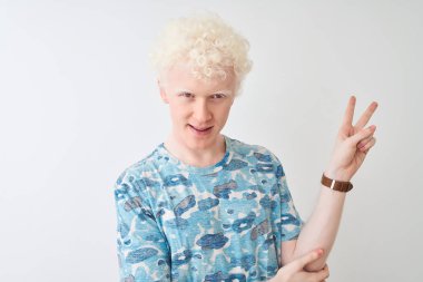 Genç albino sarışın adam rahat t-shirt izole beyaz arka plan üzerinde duran giyen mutlu yüz zafer işareti yapıyor kameraya göz kırparak gülümseyerek. İki numara.