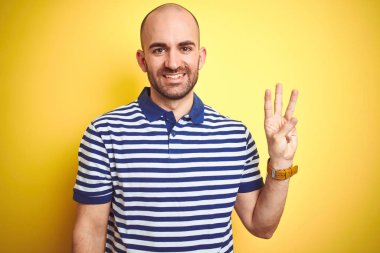 Sakallı genç ve kel bir adam sarı izole edilmiş arka plan üzerine günlük mavi tişört giyiyor ve üç numarayı gösterirken kendinden emin ve mutlu gülümsüyor..