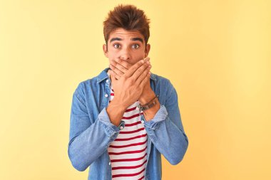 İzole edilmiş sarı arka plan üzerinde çizgili tişört ve kot gömlek giyen genç yakışıklı adam yanlışlıkla ağızlarını kapatarak şok etti. Gizli konsept.