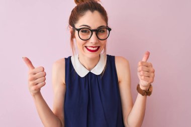 Redhead woman with pigtail wearing elegant dress and glasses over isolated pink background success sign doing positive gesture with hand, thumbs up smiling and happy. Cheerful expression and winner gesture.