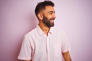 Izole pembe arka plan üzerinde duran rahat gömlek giyen genç hintli adam yüzünde gülümseme, doğal ifade ile yan bakıyor. Gülmekten emin.