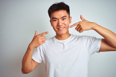 Asyalı genç bir Çinli tişört giyiyor. İzole edilmiş beyaz arka plan üzerinde duruyor. Gülümsüyor ve parmaklarını ve ağzını göstererek gülümsüyor. Diş sağlığı konsepti.