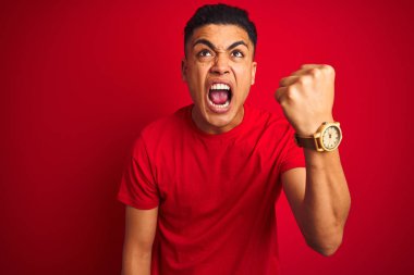 İzole edilmiş kırmızı arka plan üzerinde dikilen Brezilyalı genç adam öfkeyle bağırırken kızgın ve öfkeli yumruğunu kaldırıyor. Öfke ve agresif kavram.