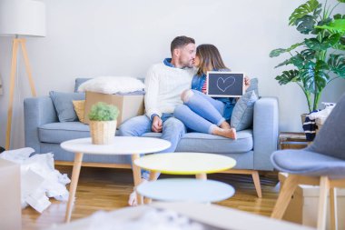 Kanepede oturan genç ve güzel bir çift ellerinde bir kalple karton kutuların etrafındaki yeni evde öpüşüyorlar.