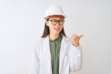 Young chinese engineer woman wearing coat helmet glasses over isolated white background smiling with happy face looking and pointing to the side with thumb up.