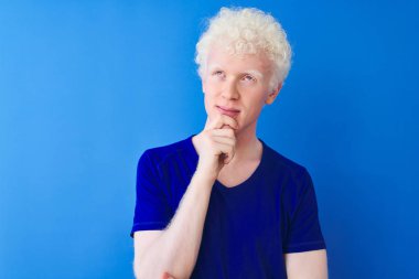 Sıradan tişörtler giyen genç albino bir adam. Mavi arka planda duruyor. Elleri çenesinde soru sorarken, düşünceli bir ifade takınıyor. Düşünceli bir yüzle gülümsüyor. Şüphe kavramı.