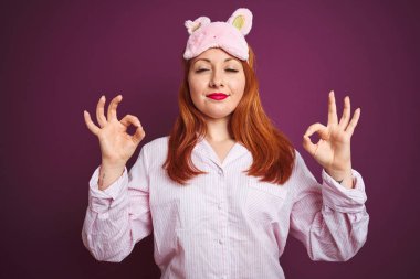 Genç, güzel, kızıl saçlı, pijama giyen ve mor izole edilmiş arka planda maske takan, gözleri kapalı, parmaklarıyla meditasyon hareketi yapan bir kadın. Yoga konsepti.