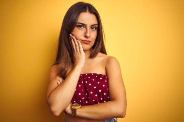 Sarı arka planda duran kırmızı tişörtlü genç ve güzel bir kadın kollarını kavuşturarak depresyondan sıkılmış ve yorgun görünüyor..