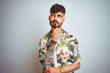 Tatilde dövmeli bir adam, izole edilmiş beyaz arka plan üzerinde dururken, elleri çenesinde soru sorarken, düşünceli bir yüz ifadesi vardı. Düşünceli bir yüzle gülümsüyor. Şüphe kavramı.