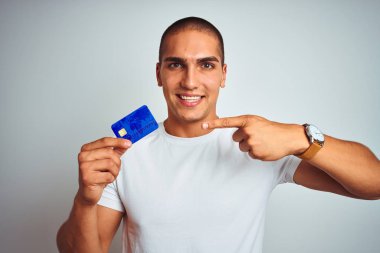 Genç ve yakışıklı bir adam kredi kartını soyutlanmış beyaz arka planda tutarken mutlu bir şekilde el ve parmağı işaret ediyor.